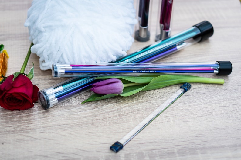 Pu&ograve; includere: Un set di ferri da maglia colorati in una custodia di plastica trasparente con un tappo nero. La custodia ha la scritta "Happy Birthday" stampata. I ferri sono accanto a un gomitolo di lana bianco, una rosa rossa e un tulipano viola.