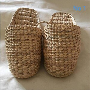 May include: A pair of woven natural fiber slippers. The slippers are light brown with a textured surface and a curved back. The slippers are made of a woven material and have a loop handle. The text "No 1" is visible in the upper right corner.