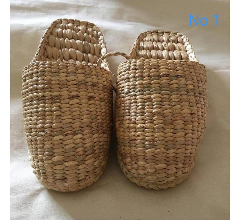 May include: A pair of woven natural fiber slippers. The slippers are light brown with a textured surface and a curved back. The slippers are made of a woven material and have a loop handle. The text "No 1" is visible in the upper right corner.