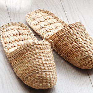 May include: Two woven, light brown slippers are displayed on a light-colored wooden floor. The slippers are made of a natural fiber, with a textured, basket-weave design. The open-toe design and neutral color suggest a comfortable, casual style.