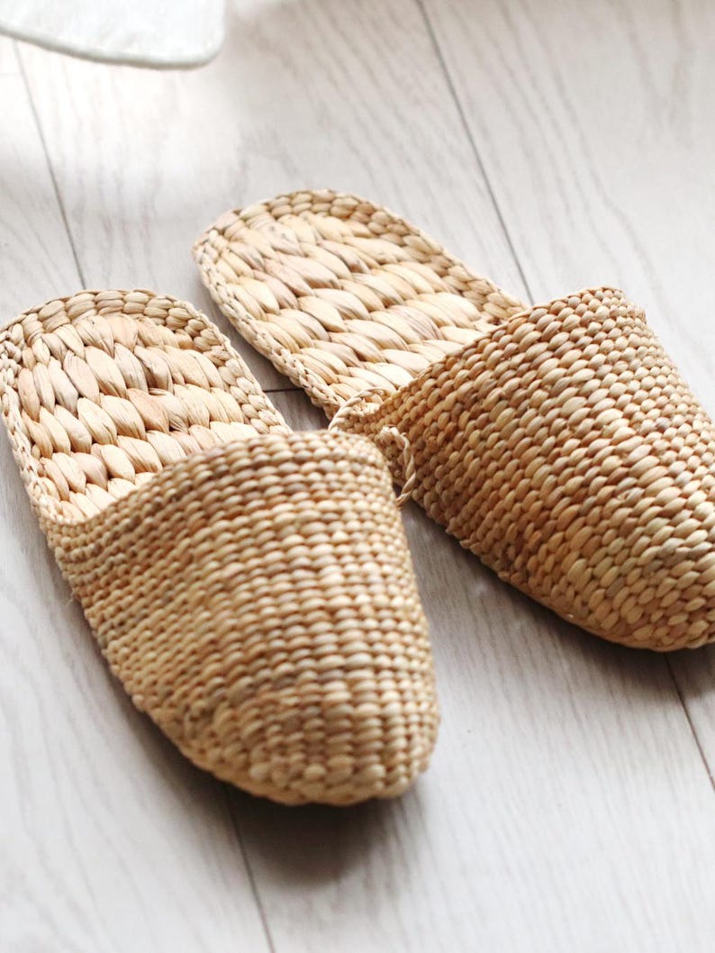 May include: Two woven, light brown slippers are displayed on a light-colored wooden floor. The slippers are made of a natural fiber, with a textured, basket-weave design. The open-toe design and neutral color suggest a comfortable, casual style.