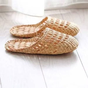May include: A pair of woven straw slippers, in a natural beige color, are displayed on a light-colored wooden surface. The slippers have an open-toe design with a textured weave pattern. The background is softly blurred, suggesting a bright, indoor setting.