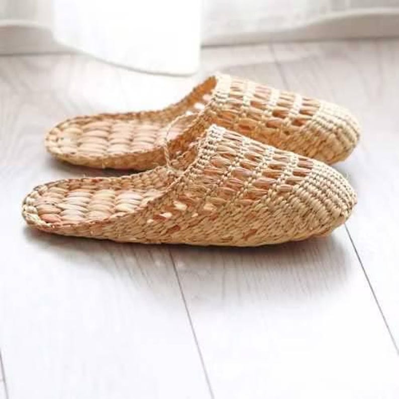 May include: A pair of woven straw slippers, in a natural beige color, are displayed on a light-colored wooden surface. The slippers have an open-toe design with a textured weave pattern. The background is softly blurred, suggesting a bright, indoor setting.