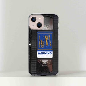 May include: A black and clear phone case designed to look like a VHS cassette tape. The case has a blue label that says "BLOCKBUSTER VIDEO" and "To PLAY is human. To REWIND is divine!" with a warning label that says "WARNING! If this label is broken, you must purchase this video."