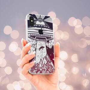 Op de afbeelding: Een witte iPhone-hoes met een zwart-witte illustratie van een UFO dat een persoon ontvoert. De achtergrond is een verloop van paars en rood.