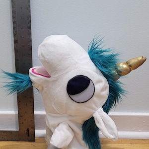 May include: A plush unicorn puppet with a white body, teal mane and tail, and a gold horn. The puppet has a dark blue eye and a pink mouth. The puppet is approximately 25 cm tall.