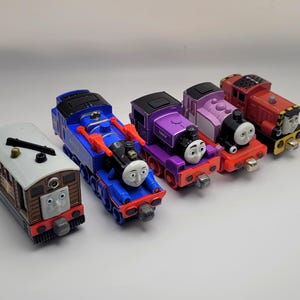 May include: A selection of toy train engines in different colours. The engines include a brown and tan carriage, a blue engine, a purple engine, a pink engine, and a red engine. Each engine has a smiling face.