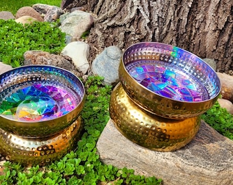 May include: Two gold metal bowls with iridescent rainbow designs inside. The bowls are sitting on rocks in a garden setting.