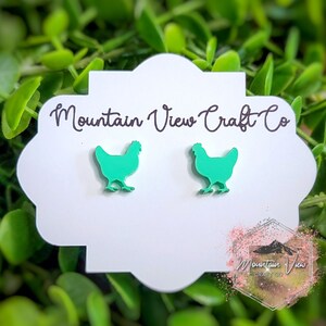 May include: A pair of bright green chicken-shaped earrings. The earrings are displayed on a white, scalloped card with the text "Mountain View Craft Co." The background is a lush green foliage.