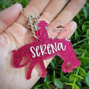 May include: Pink glitter horse keychain with a silver key ring and a pink tassel. The horse is riding a rider and the name "Serena" is written on the horse.