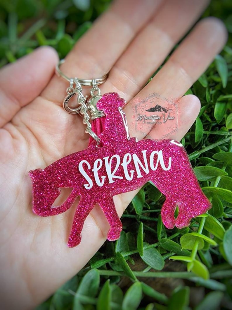 May include: Pink glitter horse keychain with a silver key ring and a pink tassel. The horse is riding a rider and the name "Serena" is written on the horse.