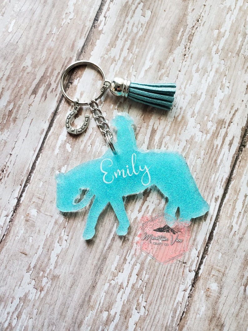 May include: A turquoise blue acrylic keychain shaped like a horse rider with the name "Emily" written on the horse's side. The keychain has a silver horseshoe charm, a silver tassel, and a silver key ring.
