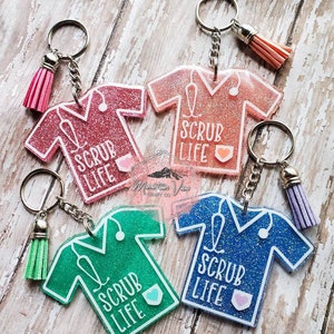 May include: Four keychains featuring a scrub top design with the text "Scrub Life" and a small heart. Each keychain has a different colour scrub top: pink, green, blue, and light blue. Each keychain has a small tassel.