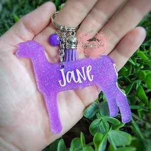 May include: Purple glitter keychain shaped like a cow with the name "Jane" written on it. The keychain has a silver charm with a purple tassel.