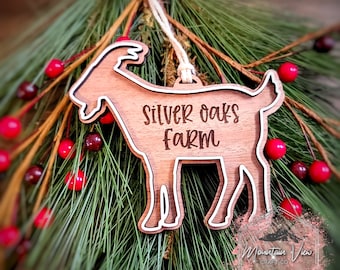 May include: Wooden goat ornament with the text "Silver Oaks Farm" in a rustic font. The ornament is brown with a white outline and a string for hanging. The ornament is displayed on a bed of green pine needles and red berries.