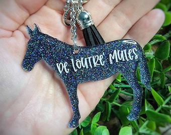 May include: A black glittery donkey-shaped keychain featuring the white text "DE LOUTRE MULES" printed on its side. The keychain includes a silver metal ring and a short black tassel attached to the top, creating a decorative accessory.