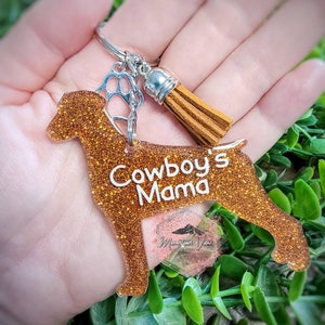 May include: A bronze glitter dog-shaped keyring with the text "Cowboy's Mama" in white. It features a paw print charm, a brown tassel, and a silver ring. A small logo is visible on the bottom.