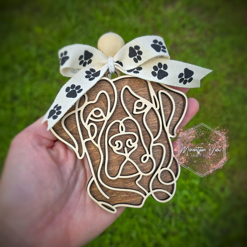 May include: Wooden ornament of a dog's face with a brown wood stain. The ornament is decorated with a white ribbon with black paw prints.