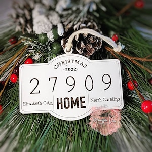 Op de afbeelding: Wit houten kerst ornament in de vorm van een bord met de tekst "Christmas -2022- 27909 Elizabeth City HOME North Carolina".