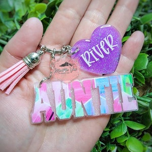 May include: A pink and blue glitter keychain with the word "AUNTIE" and a purple glitter heart with the word "River".