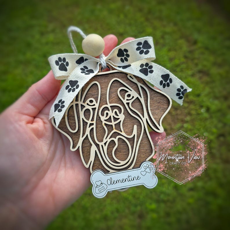 May include: Wooden dog ornament with a brown wood finish and a paw print design. The ornament is personalised with the name "Clementine" and has a white ribbon with black paw prints.