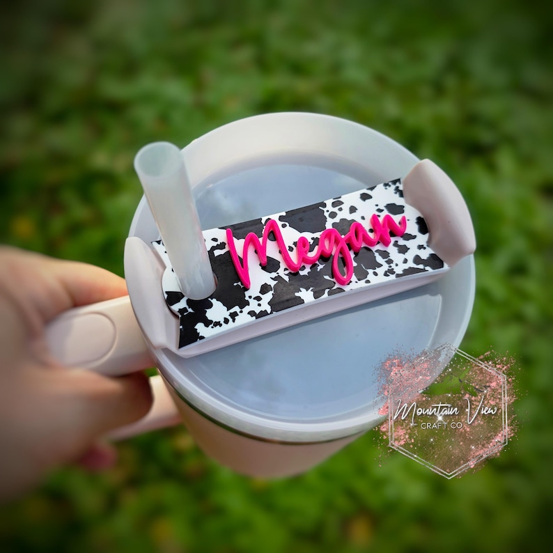 K&ouml;nnte beinhalten: Ein wei&szlig;er und schwarzer Becherdeckel mit Kuhmuster und einem rosa Namensschild mit der Aufschrift "Megan".