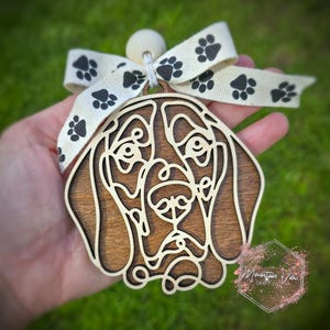 May include: A wooden ornament of a brown and white Bloodhound dog's head with a white ribbon with black paw prints. The ornament has a wooden bead at the top for hanging.