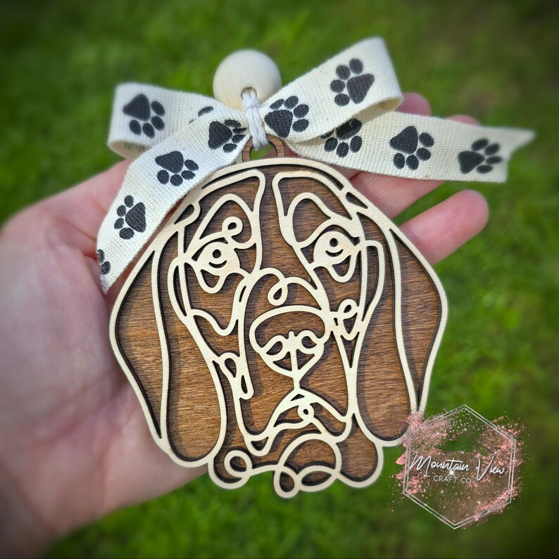 May include: A wooden ornament of a brown and white Bloodhound dog's head with a white ribbon with black paw prints. The ornament has a wooden bead at the top for hanging.