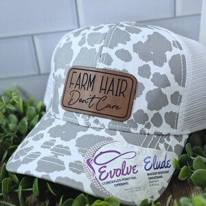 May include: A white and gray trucker hat with a cow print design. The hat features a brown leather patch with the text "FARM HAIR Don't Care." The hat has a white mesh back and a curved brim.