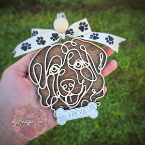 May include: Wooden dog ornament with a brown wood finish and a white bone-shaped tag with the name "FREYA" on it. The ornament is decorated with a white ribbon with black paw prints.