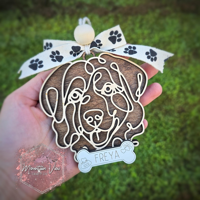 May include: Wooden dog ornament with a brown wood finish and a white bone-shaped tag with the name "FREYA" on it. The ornament is decorated with a white ribbon with black paw prints.