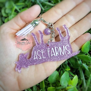 May include: Purple glitter goat keychain with a white tassel and a silver chain. The goat is upside down and the text "ASE FARMS" is printed on the goat.