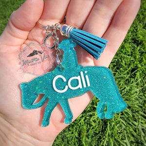 May include: Teal blue glitter horse keychain with the name "Cali" written in white. The keychain has a silver charm with a blue tassel.