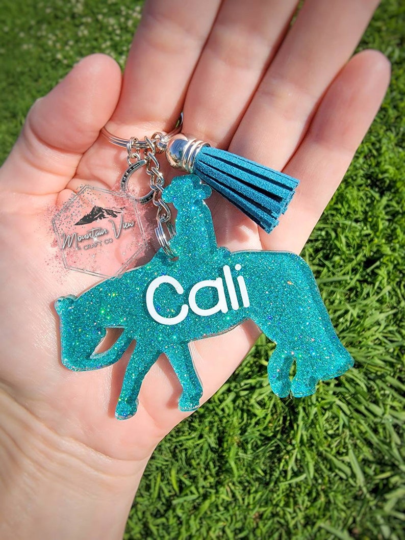 May include: Teal blue glitter horse keychain with the name "Cali" written in white. The keychain has a silver charm with a blue tassel.