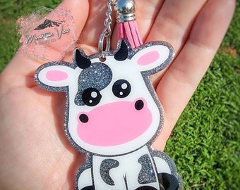 May include: A silver glitter cow keychain with black spots and a pink nose. The keychain has a silver chain and a pink tassel.