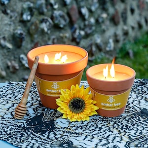 May include: Two terracotta flower pot candles with a sunflower and a wooden honey dipper on a black and white patterned surface. The candles are lit and have the text "amber bee gifts from the hive 100% Natural British Beeswax" on the label.