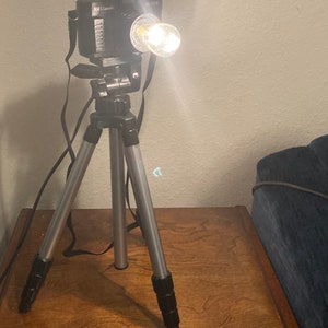 Puede incluir: Una cámara negra con una bombilla en la parte superior está montada en un trípode plateado. La cámara está sobre una mesa de madera.