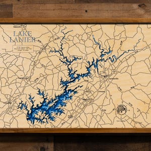 May include: Wooden framed map of Lake Lanier, with blue water detail and beige land features. The map includes roads, cities, and text that reads "Lake Lanier". The frame is a light brown color.