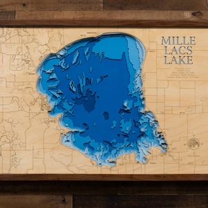 May include: A framed wooden map of Mille Lacs Lake, featuring a detailed, layered blue and light blue depiction of the lake's depth. The map is set against a light wood background with the lake's name in the upper right corner.