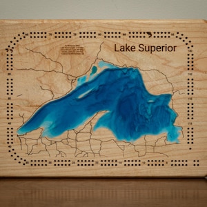 May include: A rectangular wooden cribbage board featuring a laser-engraved map of Lake Superior. The lake is filled with blue resin, and the board includes numbered scoring tracks along the perimeter with text detailing lake statistics.