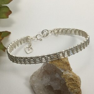 May include: A silver-colored wire-wrapped bracelet with a heart charm. The bracelet features a woven design and a clasp closure. The heart charm is a small, open design. The bracelet is displayed on a light-colored surface.