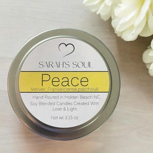 May include: A silver candle tin with a white label that reads "SARAH'S SOUL Peace Vetiver. Frankincense,patchouli Hand Poured in Holden Beach NC Soy Blended Candles Created With Love & Light Net wt 3.15 oz"