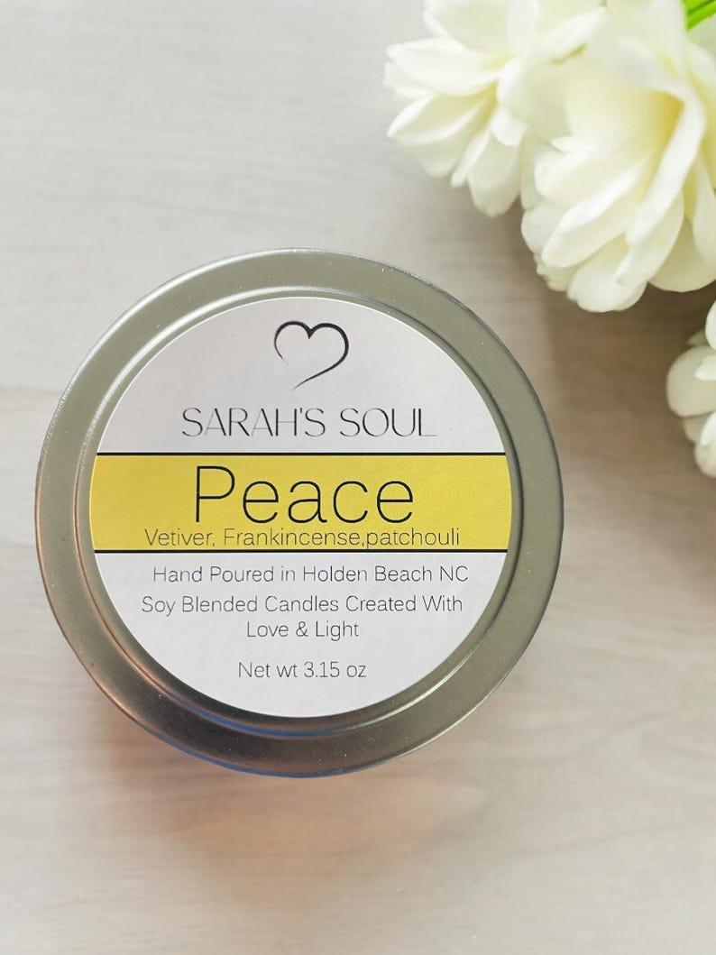 May include: A silver candle tin with a white label that reads "SARAH'S SOUL Peace Vetiver. Frankincense,patchouli Hand Poured in Holden Beach NC Soy Blended Candles Created With Love & Light Net wt 3.15 oz"