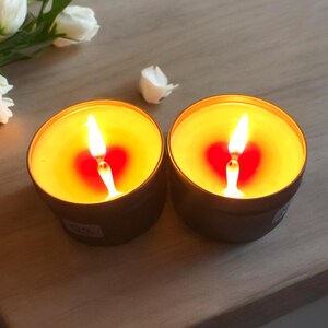 May include: Two gold-colored candle jars with a heart-shaped red wax pool and a lit wick in each jar. The jars are on a light brown wooden surface.