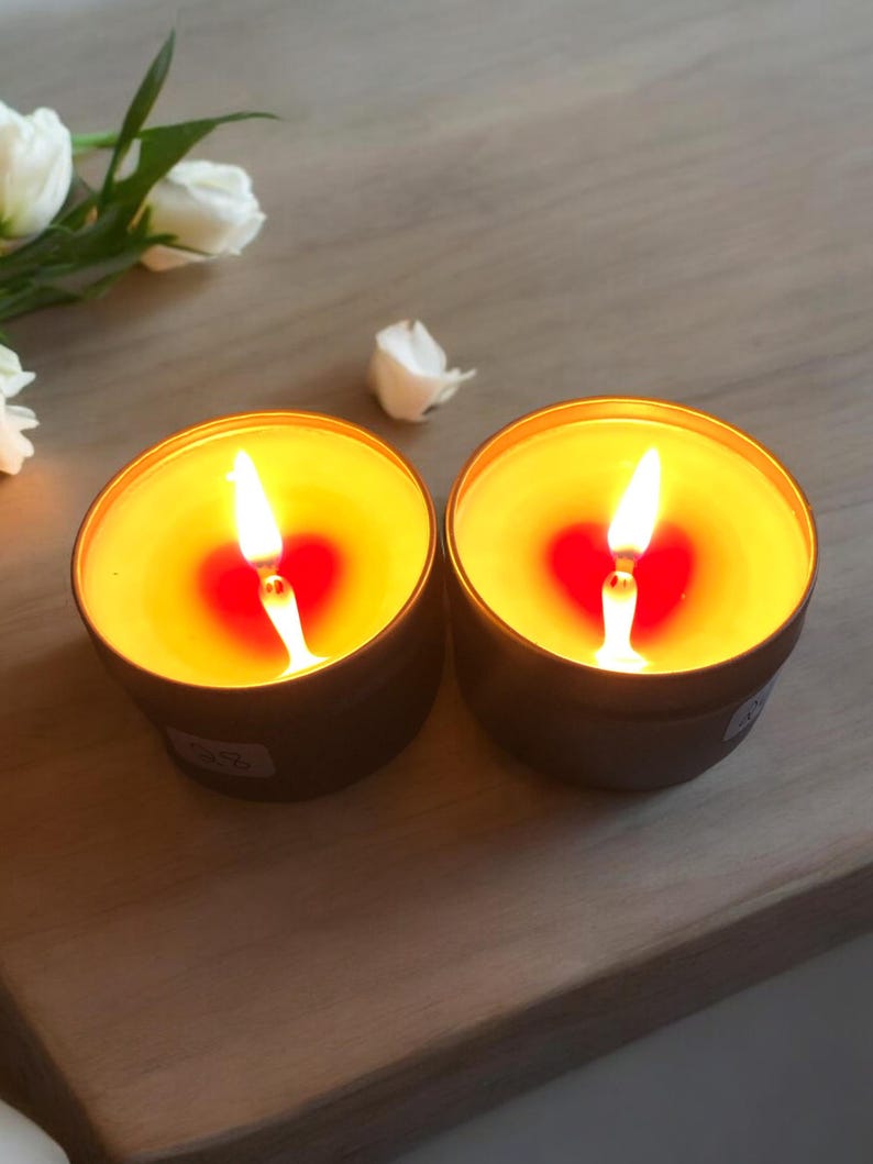 May include: Two gold-colored candle jars with a heart-shaped red wax pool and a lit wick in each jar. The jars are on a light brown wooden surface.