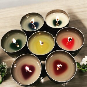 May include: Six small, round candles in metal tins with various colored waxes and flames burning. The candles are arranged in a circle on a wooden surface.