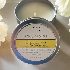 May include: A silver tin candle with a white label that reads "SARAH'S SOUL Peace Vetiver, Frankincense, patchouli Hand Poured in Holden Beach NC Soy Blended Candles Created With Love & Light Net wt 3.15 oz".