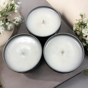 May include: Three white candles in gray metal containers. The candles are unlit and have a white wax surface.