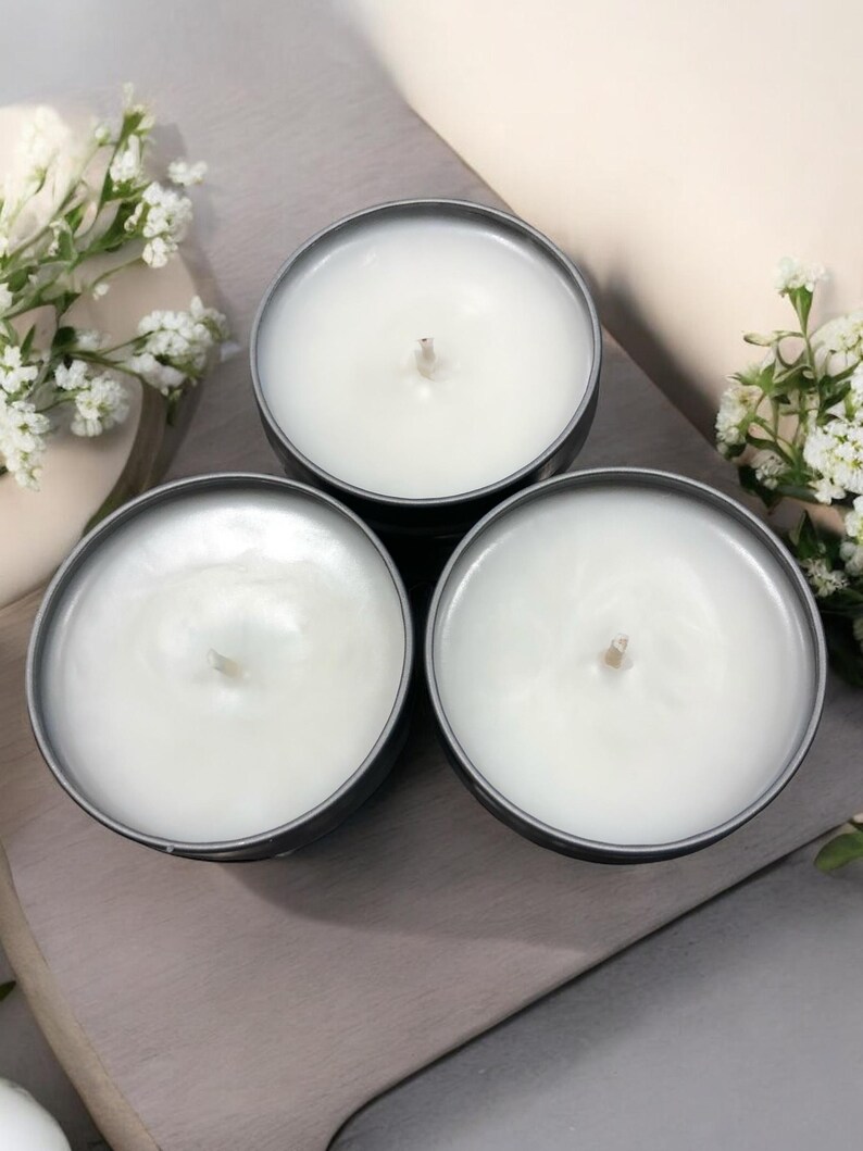 May include: Three white candles in gray metal containers. The candles are unlit and have a white wax surface.