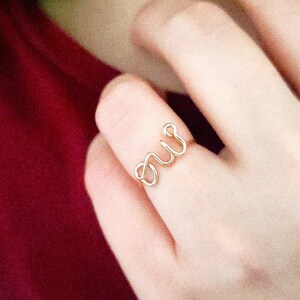Peut inclure: Une bague en fil métallique doré présentant un design en écriture cursive continue formant le mot 'oui'. La bague est fabriquée à partir d'une seule pièce de fil métallique fin et poli, créant un anneau délicat et léger sur le doigt.